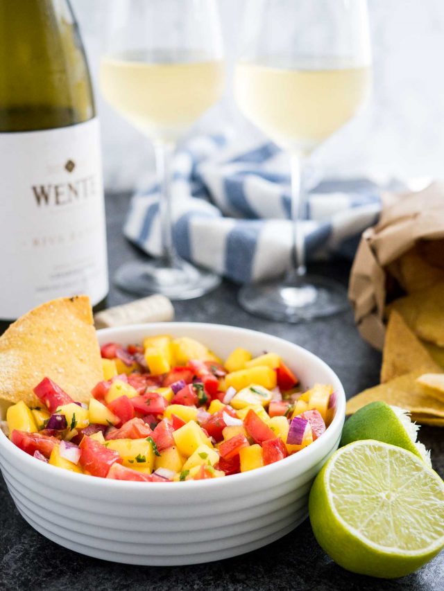 Un bol blanc contenant de la salsa aux tomates et à la mangue avec une chips à l'intérieur sur une surface sombre à côté de deux citrons verts, un sachet de nachos et un torchon blanc et bleu. Derrière, il y a une bouteille et deux verres de vin blanc.