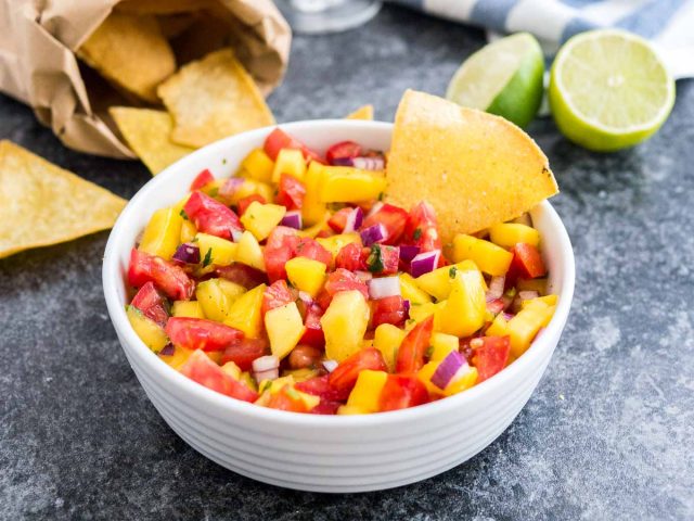 Un bol blanc contenant de la salsa aux tomates et à la mangue avec une chips à l'intérieur sur une surface sombre à côté de deux citrons verts, un sachet de nachos et un torchon blanc et bleu.
