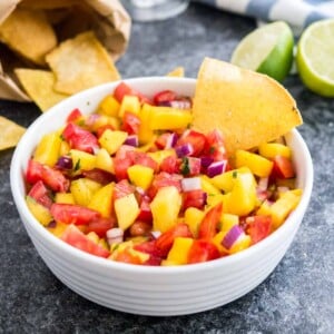 Un bol blanc contenant de la salsa aux tomates et à la mangue avec une chips à l'intérieur sur une surface sombre à côté de deux citrons verts, un sachet de nachos et un torchon blanc et bleu.
