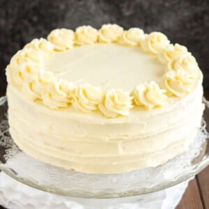 Un plat de service en verre avec un gâteau au citron et aux graines de pavot sur une table en bois avec une nappe blanche.