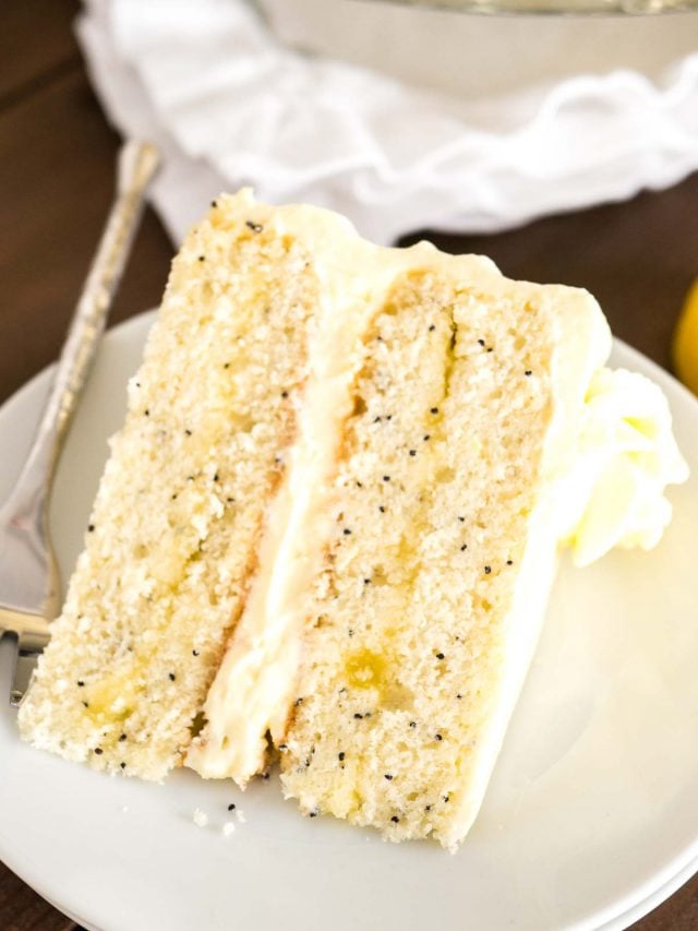 Une part de gâteau au citron et aux graines de pavot sur une assiette blanche avec une fourchette. Il y a un torchon blanc et un citron à côté.