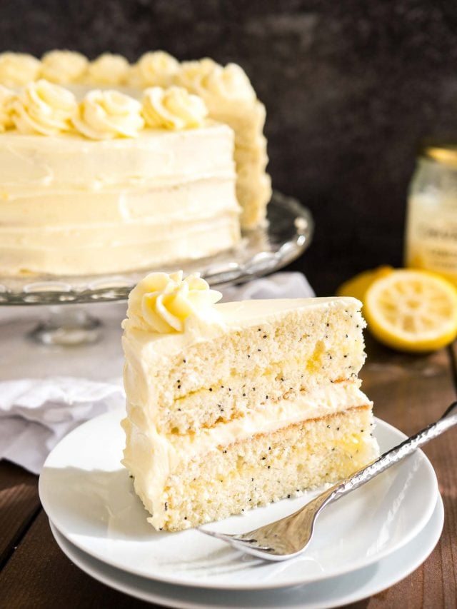Une part de gâteau au citron et aux graines de pavot sur une assiette blanche avec une fourchette. Le reste du gâteau est sur un plat de service en verre à l'arrière-plan à côté de quelques citrons.