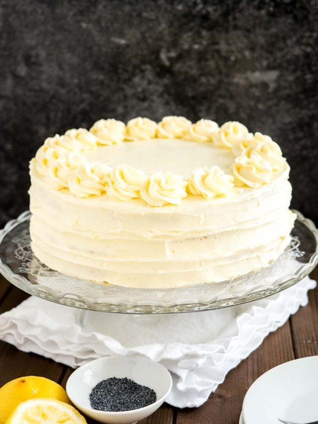 Un plat de service en verre avec un gâteau au citron et aux graines de pavot sur une table en bois avec une nappe blanche.