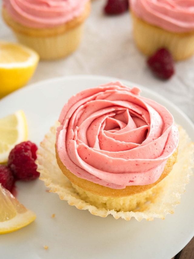 Gros plan d'un cupcake au citron et framboise avec glaçage framboise rose, avec la caissette ouverte sur une assiette blanche, garni de tranches de citron et de framboises. D'autres cupcakes se trouvent en arrière-plan.