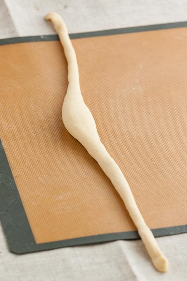 Pâte à bretzel roulée en une longue forme de saucisse avec une partie centrale épaisse sur un tapis de cuisson en silicone.