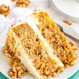Une part de gâteau aux carottes et ananas garnie de noix reposant sur le côté d'une assiette blanche empilée sur une assiette turquoise avec une fourchette.