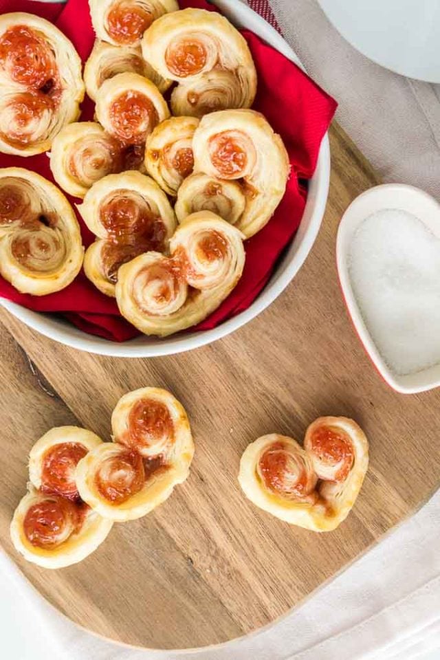 Vue de dessus de palmiers en pâte feuilletée en forme de cœur. Trois sont sur une planche à découper et le reste est dans un bol blanc recouvert d'une serviette rouge.