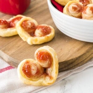 Palmiers en pâte feuilletée en forme de cœur. Un est devant appuyé contre une planche à découper en bois, les autres sont posés sur la planche et dans un bol blanc sur la planche.