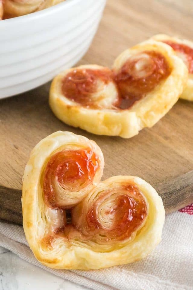 Gros plan d'un palmier en pâte feuilletée en forme de cœur appuyé contre une planche à découper en bois avec d'autres palmiers en arrière-plan.