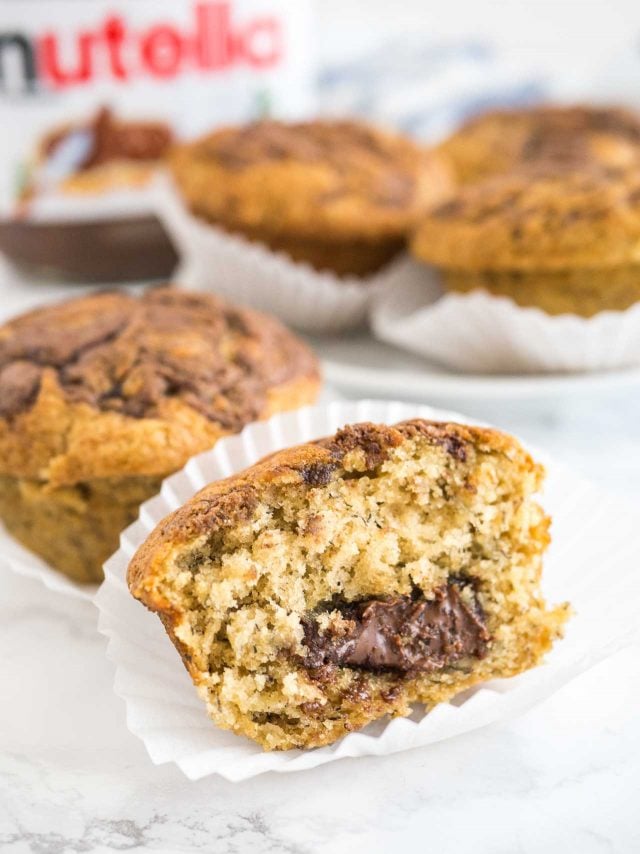 Gros plan d'un muffin banane nutella dans sa caissette avec une grosse bouchée prise. Il y a d'autres muffins et un pot de nutella en arrière-plan.