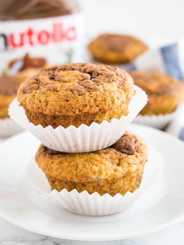 Gros plan de deux muffins banane nutella empilés l'un sur l'autre sur une assiette blanche, avec d'autres muffins et un pot de nutella en arrière-plan.