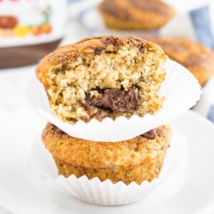 Deux muffins banane nutella empilés l'un sur l'autre sur une assiette blanche, avec d'autres muffins et un pot de nutella en arrière-plan. Le muffin du dessus a déjà une grosse bouchée.