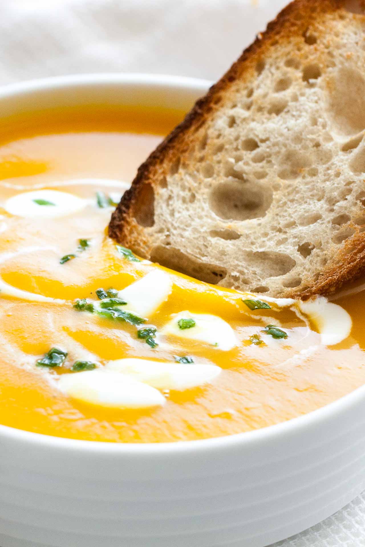 Gros plan d'une tranche de pain grillé trempée dans de la soupe de potiron butternut avec des cuillerées de crème aigre et de ciboulette dans un bol blanc.