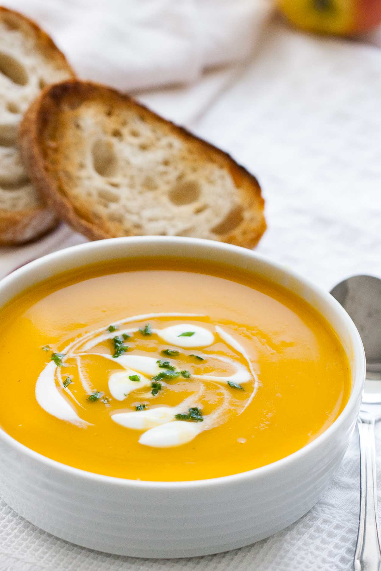 Un bol blanc avec de la soupe de potiron butternut, avec des cuillerées de crème aigre et de la ciboulette. Il est posé sur un torchon blanc et une cuillère ainsi que des tranches de pain grillé sont à côté.