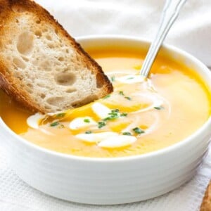 Un bol blanc avec de la soupe de potiron butternut, avec des cuillerées de crème aigre et de la ciboulette, une cuillère dedans et une tranche de pain grillé. Il est posé sur un torchon blanc à côté de quelques tranches de pain grillé.