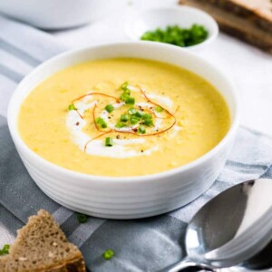 Un bol blanc de soupe crémeuse de maïs au curry, garnie de crème aigre, de fils de piment et de ciboulette sur un torchon gris. Un morceau de pain devant et une cuillère à côté. Un autre bol de soupe, un petit bol blanc de ciboulette et plus de pain en arrière-plan.