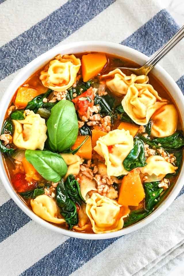 Vue de dessus d'un bol blanc de soupe de tortellini aux épinards garnie de feuilles de basilic sur un torchon blanc et bleu.