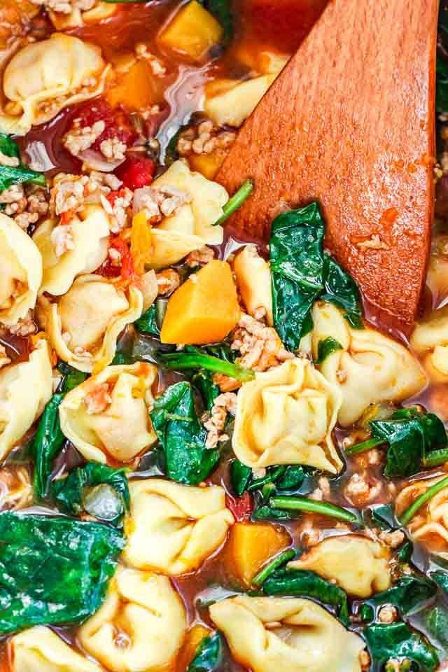 Gros plan d'un bol blanc de soupe de tortellini aux épinards garnie de feuilles de basilic avec une spatule en bois.