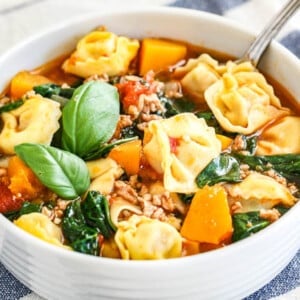 Un bol blanc de soupe de tortellini aux épinards avec une cuillère à l'intérieur et garnie de feuilles de basilic à côté d'un torchon blanc et bleu.