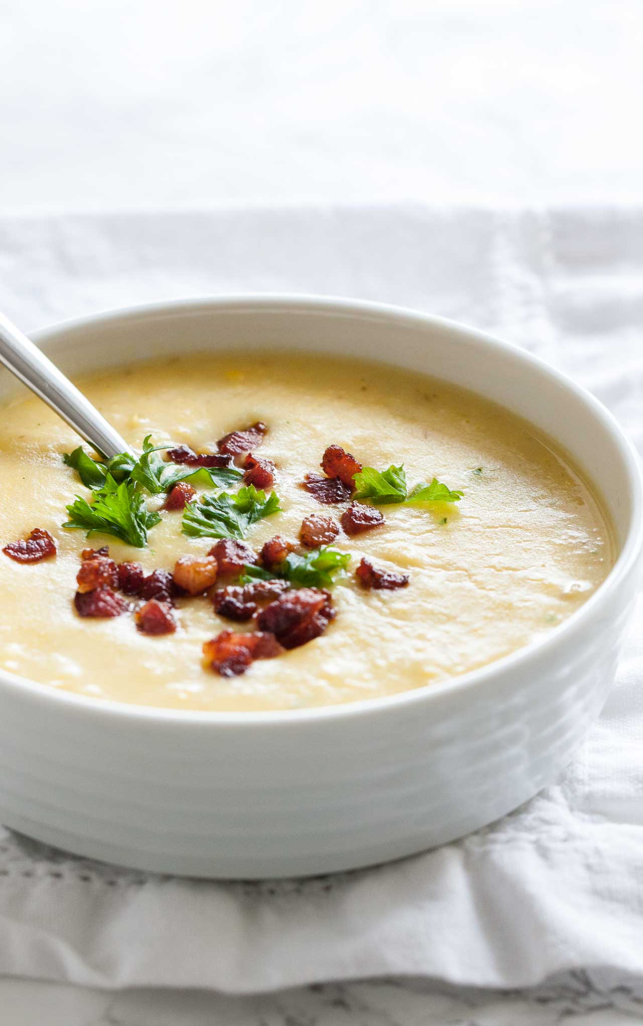 Un bol blanc de soupe de pommes de terre garni de bacon et de persil avec une cuillère dedans sur une nappe blanche.