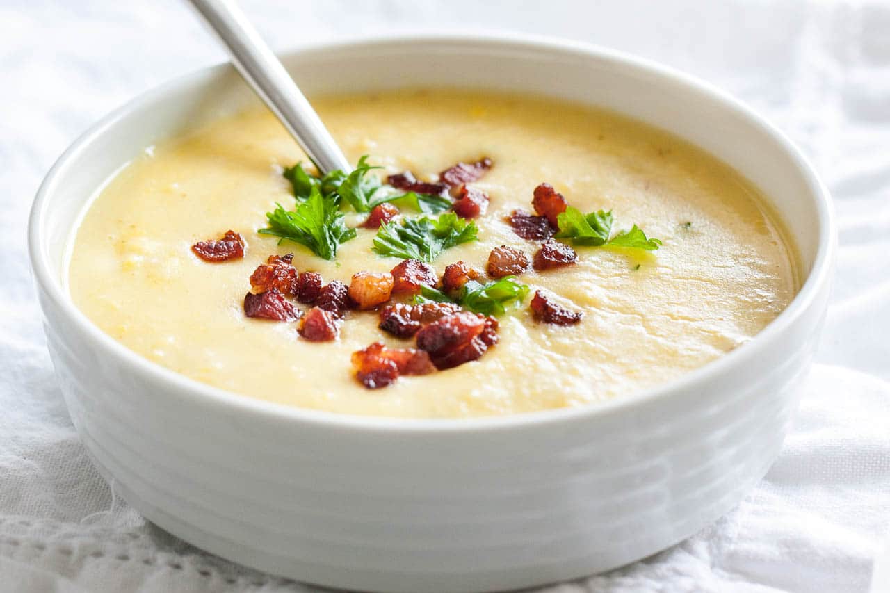 Un bol blanc de soupe de pommes de terre garni de bacon et de persil avec une cuillère dedans sur une nappe blanche.
