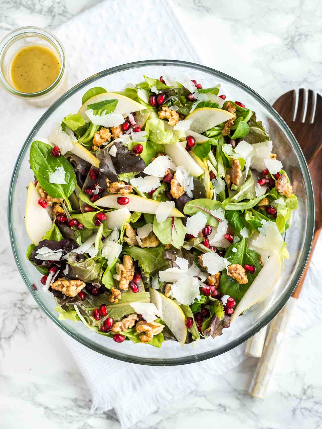 Vue de dessus d'un bol en verre avec une salade de poire et de grenade avec des noix sur un torchon blanc sur une surface en marbre avec des ustensiles de salade en bois et un petit bol de vinaigrette.