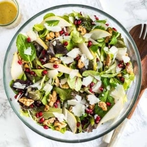 Vue de dessus d'un bol en verre avec une salade de poire et de grenade avec des noix sur un torchon blanc sur une surface en marbre avec des ustensiles de salade en bois et un petit bol de vinaigrette.