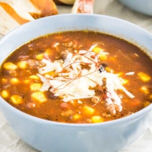 Un bol gris de chili con carne avec de la bière et du maïs, garni de fils de chili et de fromage râpé. Il y a des tranches de pains bretzel en arrière-plan.