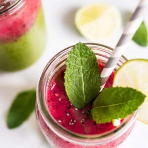 Un verre de smoothie à l'eau de coco, avec des framboises, des épinards, du chia et de la mangue, garni de menthe et d'une tranche de citron vert avec une paille. Il y a un autre verre de smoothie et des citrons verts et de la menthe à côté.