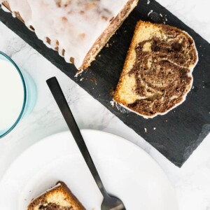 Vue de dessus d'un gâteau marbré à l'orange sur une dalle de granit gris avec une tranche à l'avant. Il y a un verre de lait et une assiette blanche avec une autre tranche ainsi qu'une fourchette noire à côté.