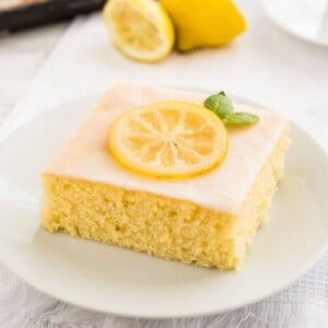 Une part de gâteau au citron avec glaçage au citron, une rondelle de citron et de la menthe sur une assiette blanche sur un torchon blanc. Il y a deux moitiés de citron, une tasse en porcelaine blanche et le reste du gâteau rectangulaire en arrière-plan, avec une fourchette à côté.
