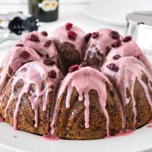 Gâteau bundt au chocolat et aux canneberges avec un glaçage rose, garni de canneberges séchées sur une assiette blanche. En arrière-plan, il y a un sucrier en acier inoxydable et un tire-bouchon.