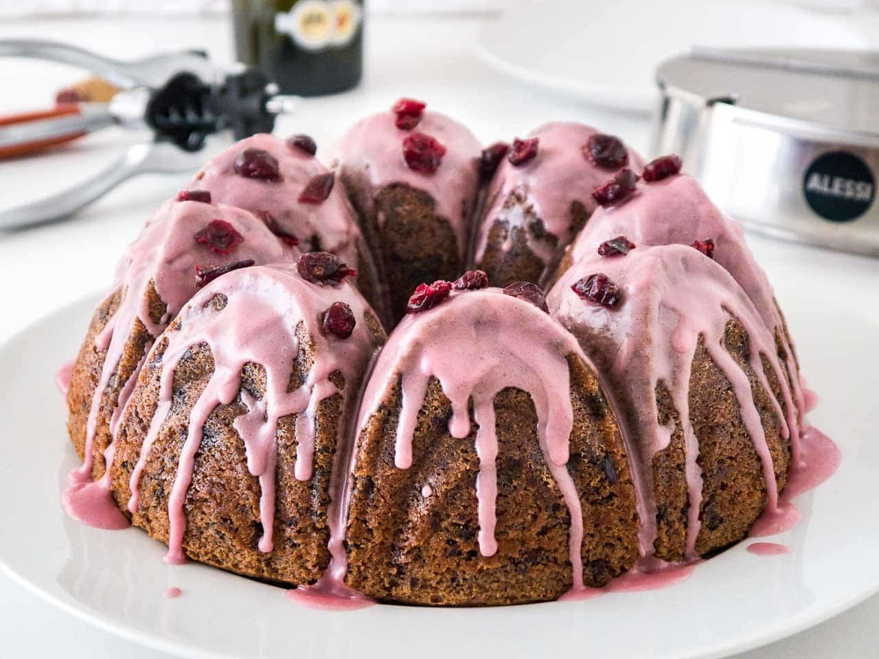 Gâteau bundt au chocolat et aux canneberges avec un glaçage rose, garni de canneberges séchées sur une assiette blanche. En arrière-plan, il y a un sucrier en acier inoxydable et un tire-bouchon.