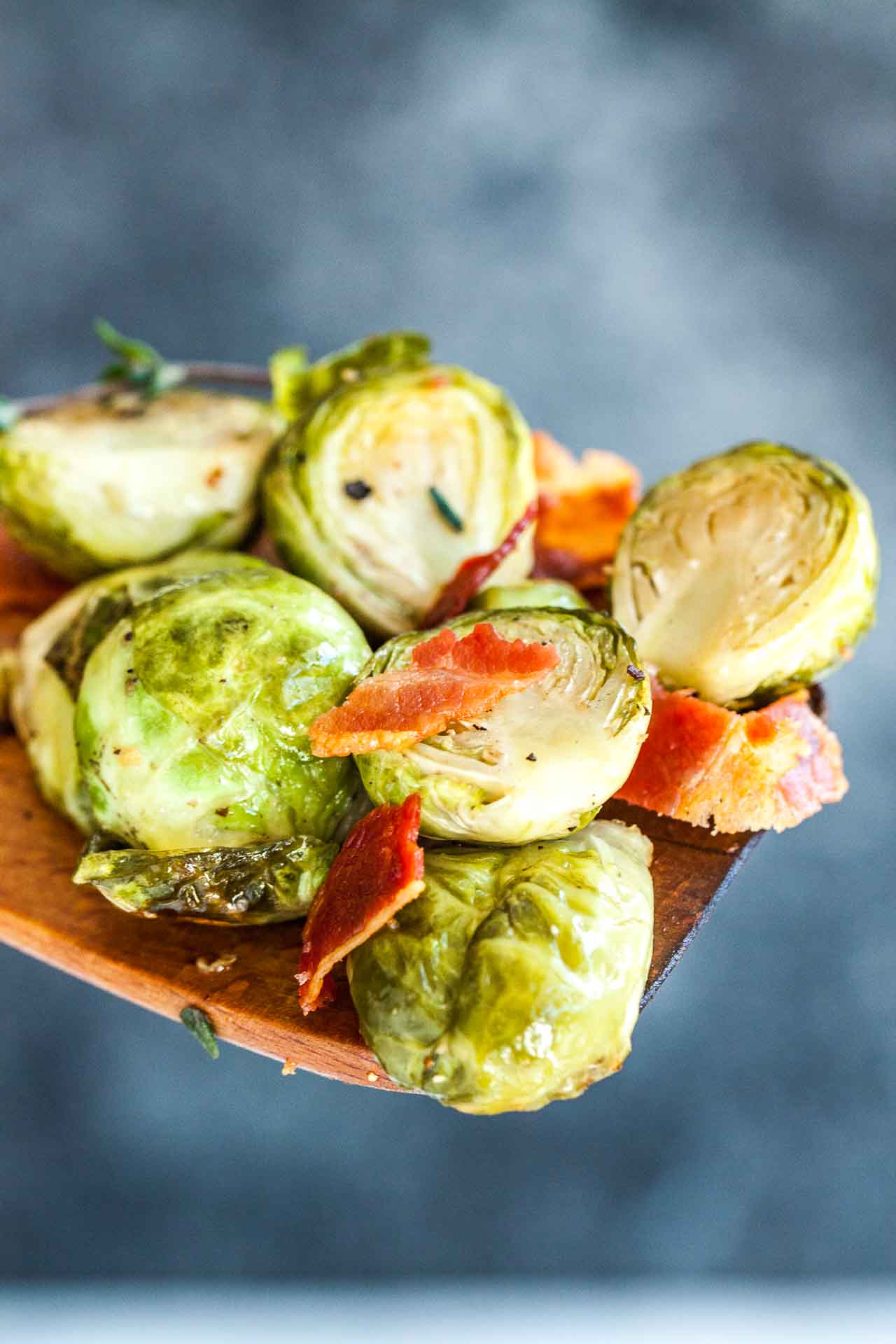 Choux de Bruxelles rôtis avec du bacon sur une spatule en bois devant un fond gris.