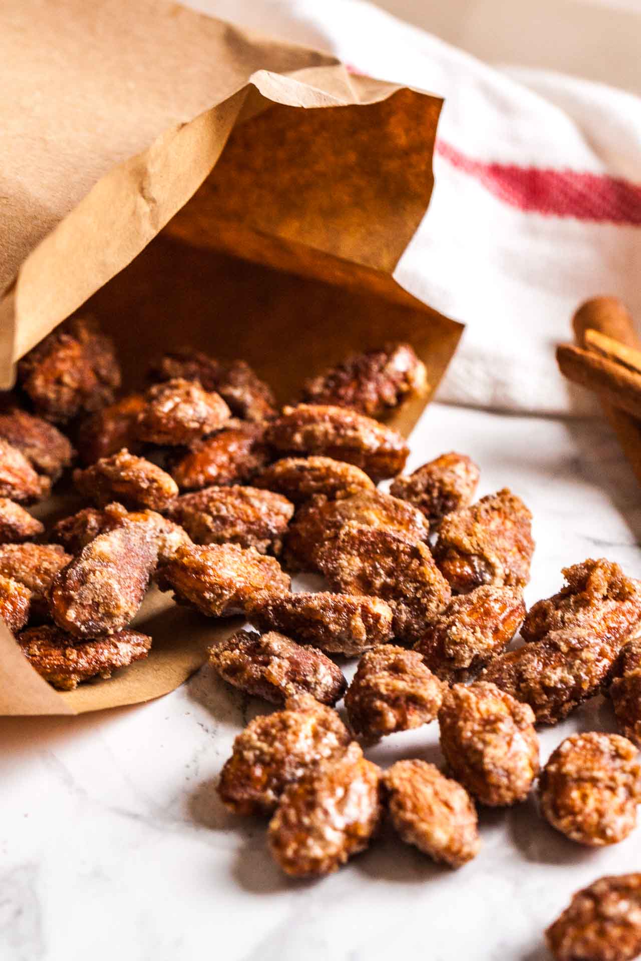Un sac en papier brun, couché sur le côté avec des amandes caramélisées à la cannelle qui débordent sur une surface en marbre, garnies de bâtons de cannelle et d'un torchon blanc et rouge en arrière-plan.