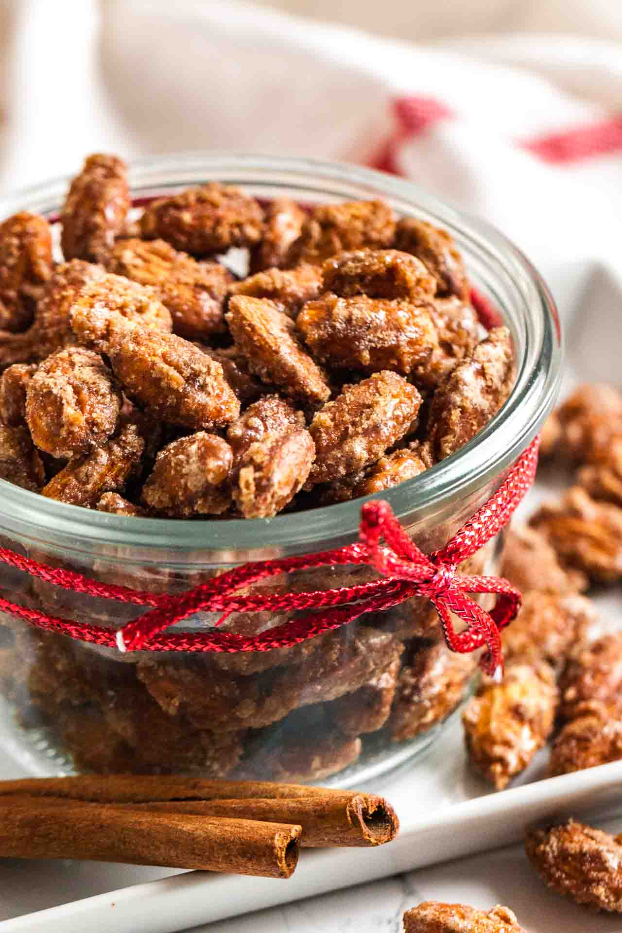 Un bol en verre d'amandes caramélisées à la cannelle avec un ruban rouge autour à côté d'un peu plus d'amandes.