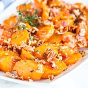 Une assiette rectangulaire blanche de carottes glacées au sucre brun avec du bacon et des noix de pécan garnie de thym.