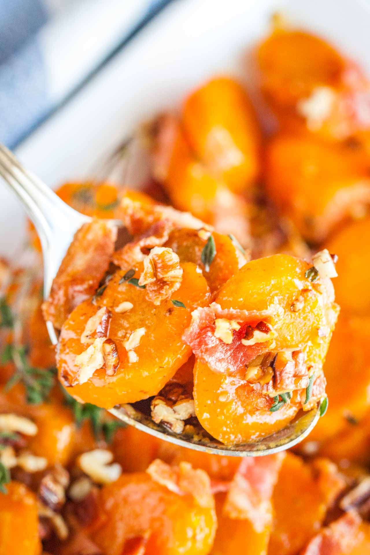 Une cuillère tenant des carottes glacées au sucre brun avec du bacon et des noix de pécan au-dessus d'un bol blanc de carottes.