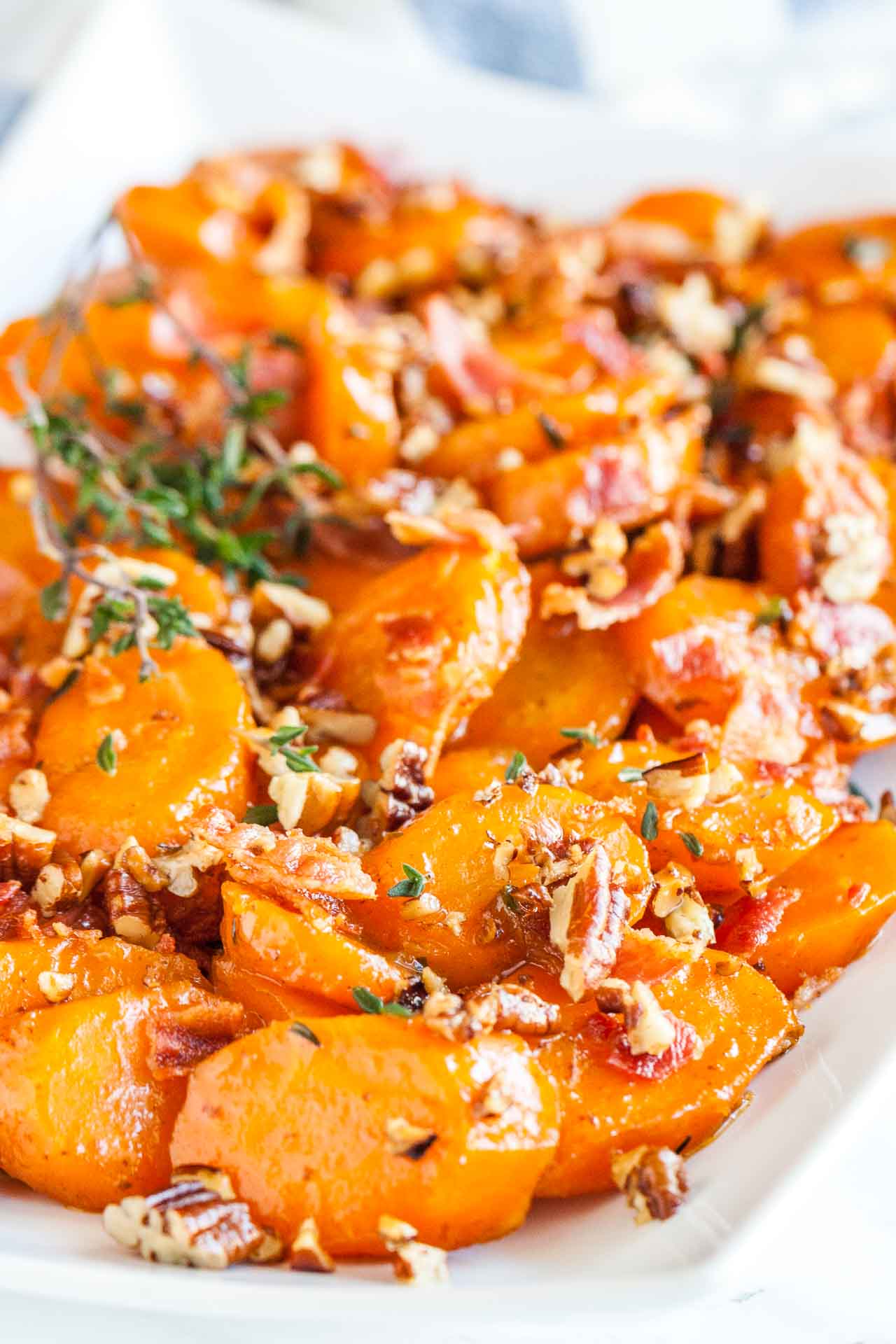 Gros plan d'une assiette rectangulaire blanche de carottes glacées au sucre brun avec du bacon et des noix de pécan garnie de thym.