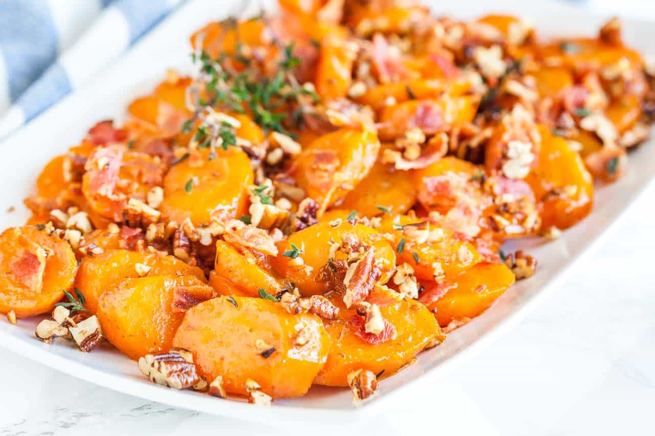 Une assiette rectangulaire blanche de carottes glacées au sucre brun avec du bacon et des noix de pécan garnie de thym.