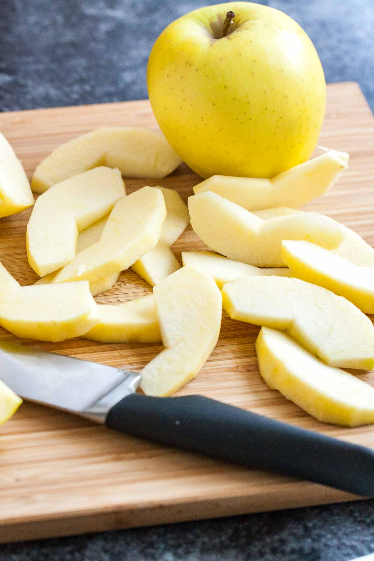 Une planche à découper en bambou avec des quartiers de pommes épluchées, une pomme entière et un couteau.
