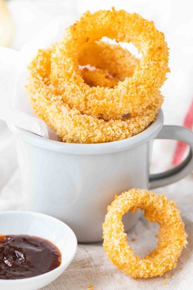 Rondelles d'oignon frites dans une tasse grise avec du papier parchemin. Il y a un petit bol en porcelaine blanche avec de la sauce barbecue à côté et une rondelle d'oignon seule se penchant contre elle.