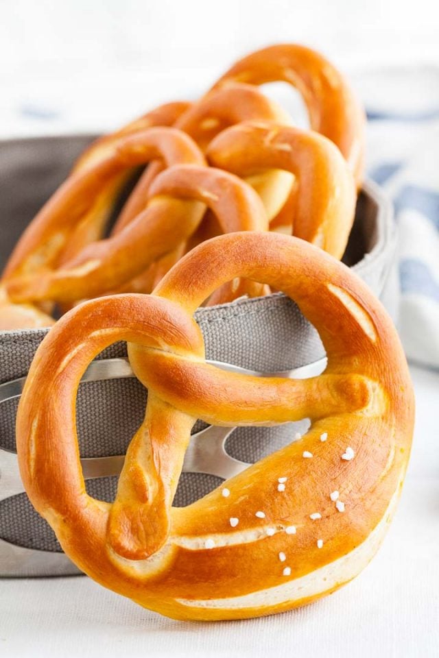 Un bretzel appuyé contre un panier à pain, contenant d'autres bretzels avec un torchon blanc et bleu en arrière-plan.