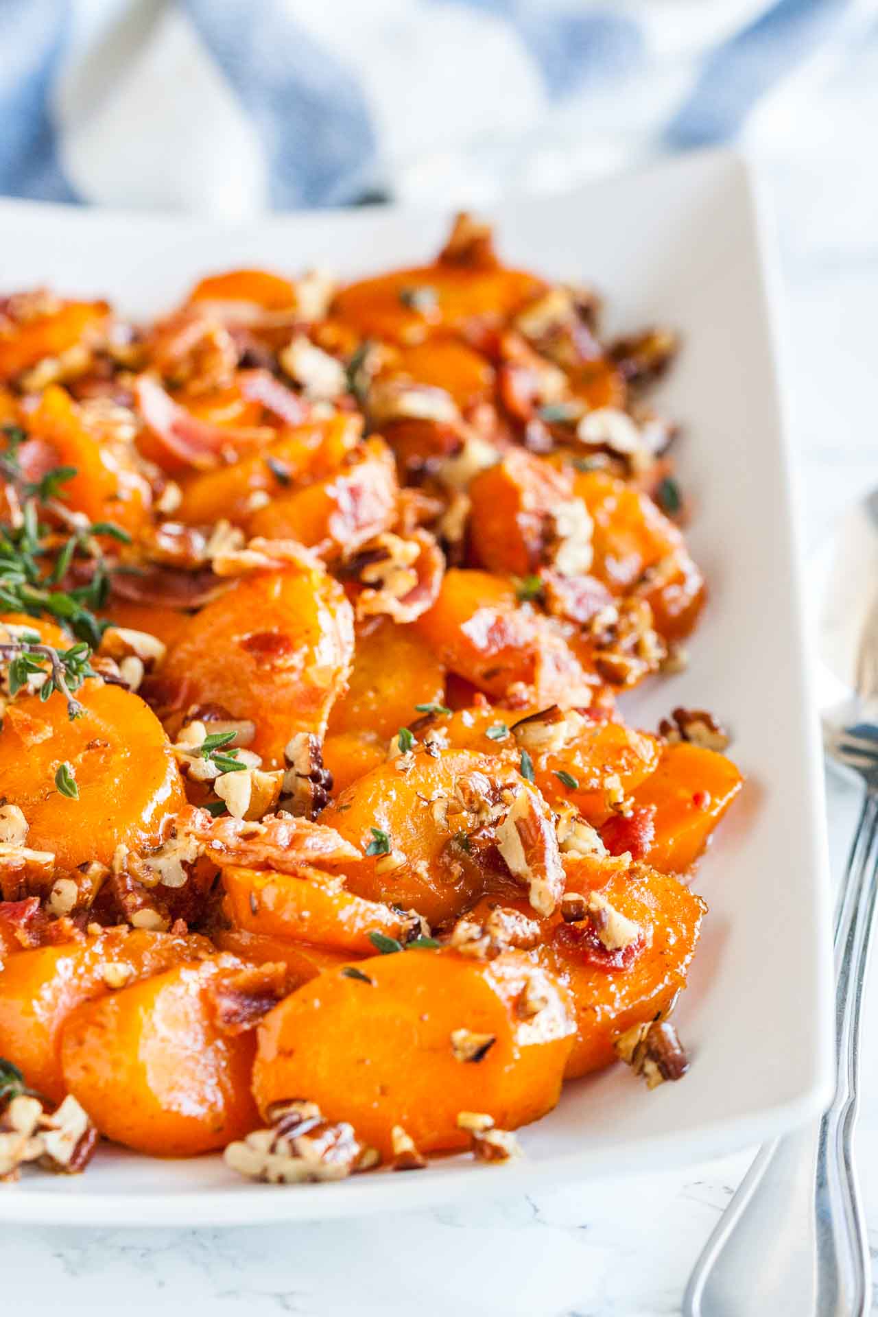 Une assiette rectangulaire blanche de carottes glacées au sucre brun avec du bacon et des noix de pécan garnie de thym sur une surface en marbre avec une cuillère à côté.