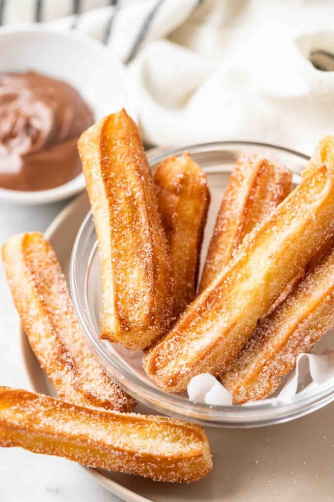 churros sucrés dans un bol en verre avec du papier sulfurisé, avec un petit bol blanc de nutella en arrière-plan