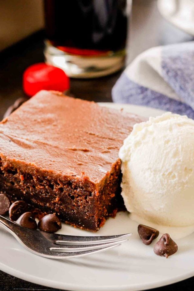 une tranche de gâteau au coca-cola sur une assiette blanche avec de la glace à la vanille, une fourchette et des pépites de chocolat, avec un torchon blanc et bleu et un bouchon de bouteille de coca-cola en arrière-plan
