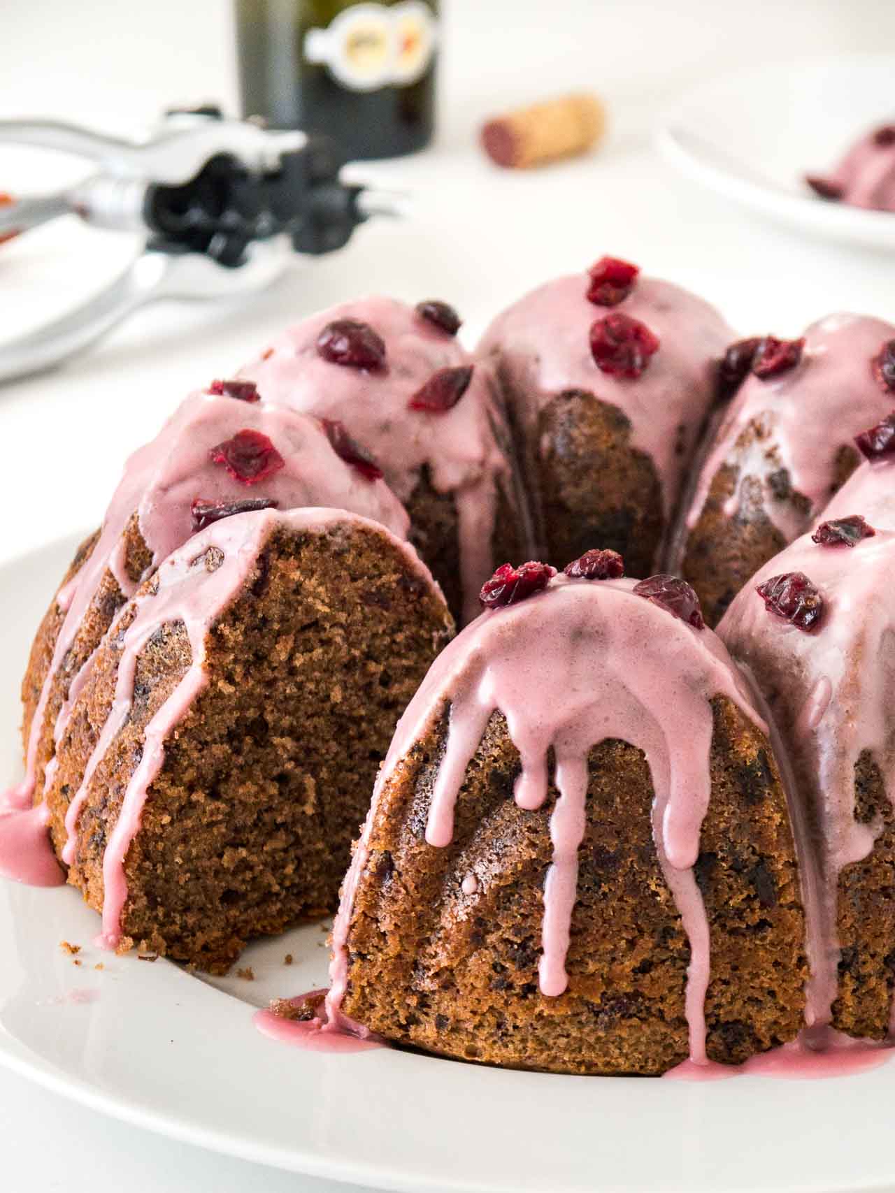 Gâteau bundt au chocolat et aux canneberges avec un glaçage rose, garni de canneberges séchées sur une assiette blanche. Un morceau du gâteau a été découpé. En arrière-plan, il y a un sucrier en acier inoxydable et un tire-bouchon.