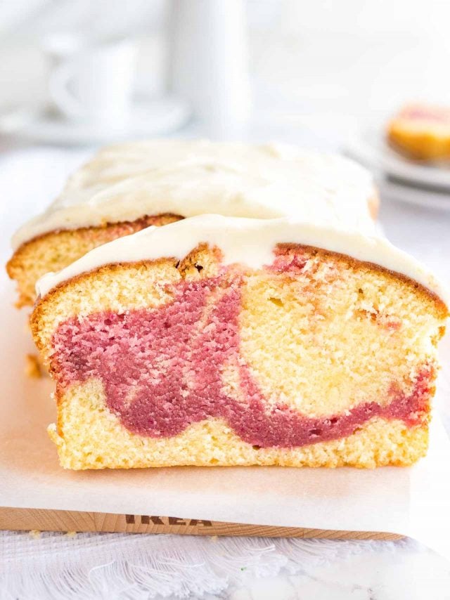 Un gâteau au yaourt aux framboises sur une planche à découper en bois recouverte de papier sulfurisé. Une tranche a été coupée et est appuyée contre. En arrière-plan, il y a deux assiettes avec une autre tranche.