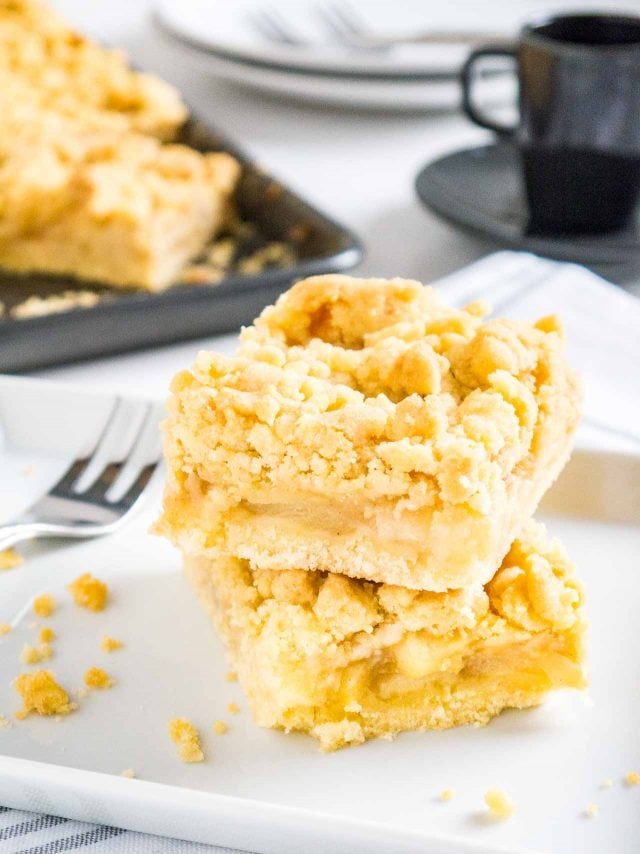 deux morceaux de gâteau streusel aux pommes empilés l'un sur l'autre sur du papier sulfurisé avec une fourchette à côté et une plaque de cuisson en arrière-plan