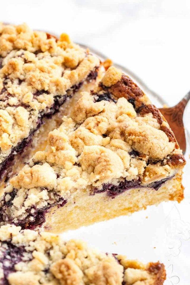 Un gâteau aux myrtilles entier avec garniture streusel sur un plat de service en verre. Un serveur à gâteau soulève une part.